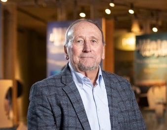 A man wears a dark blue/grey plaid suit jacket and light blue button down shirt.