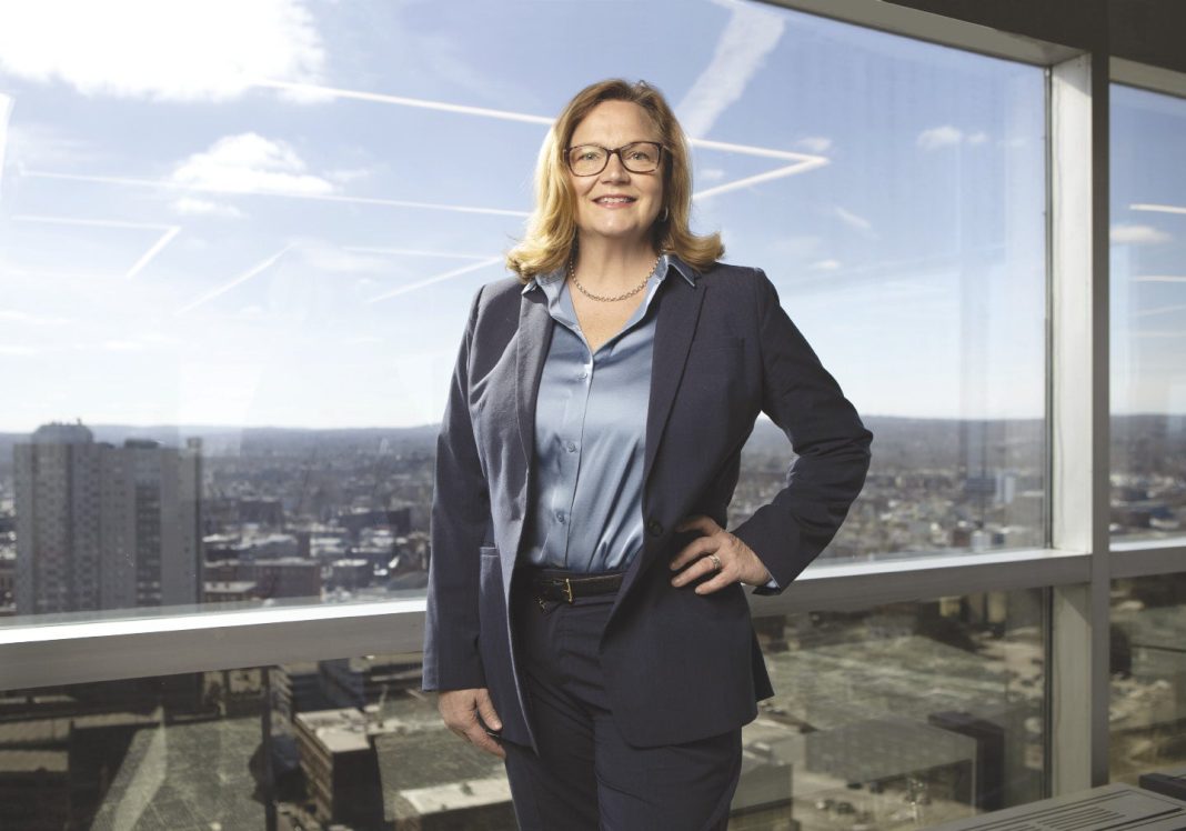 A woman in a suit jacket stands in front of a window overlooking the Worcester skyline
