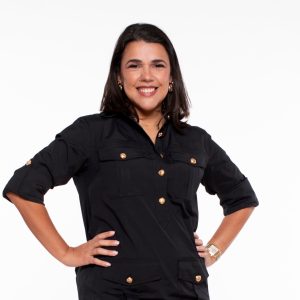 A woman with shoulder length dark hair wears a black dress with gold buttons with her hands on her hips.