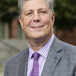 A man with gray hair wears a gray suit with a purple tie