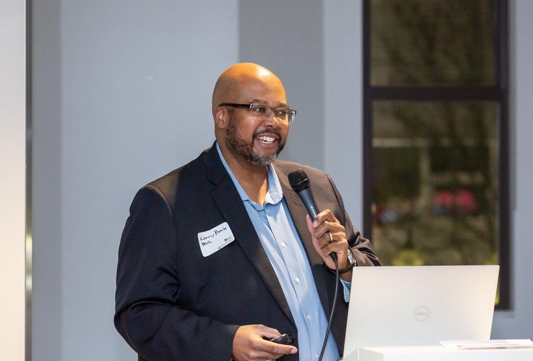 A man with glasses in a black suit jacket and light blue shirt speaks into a microphone.