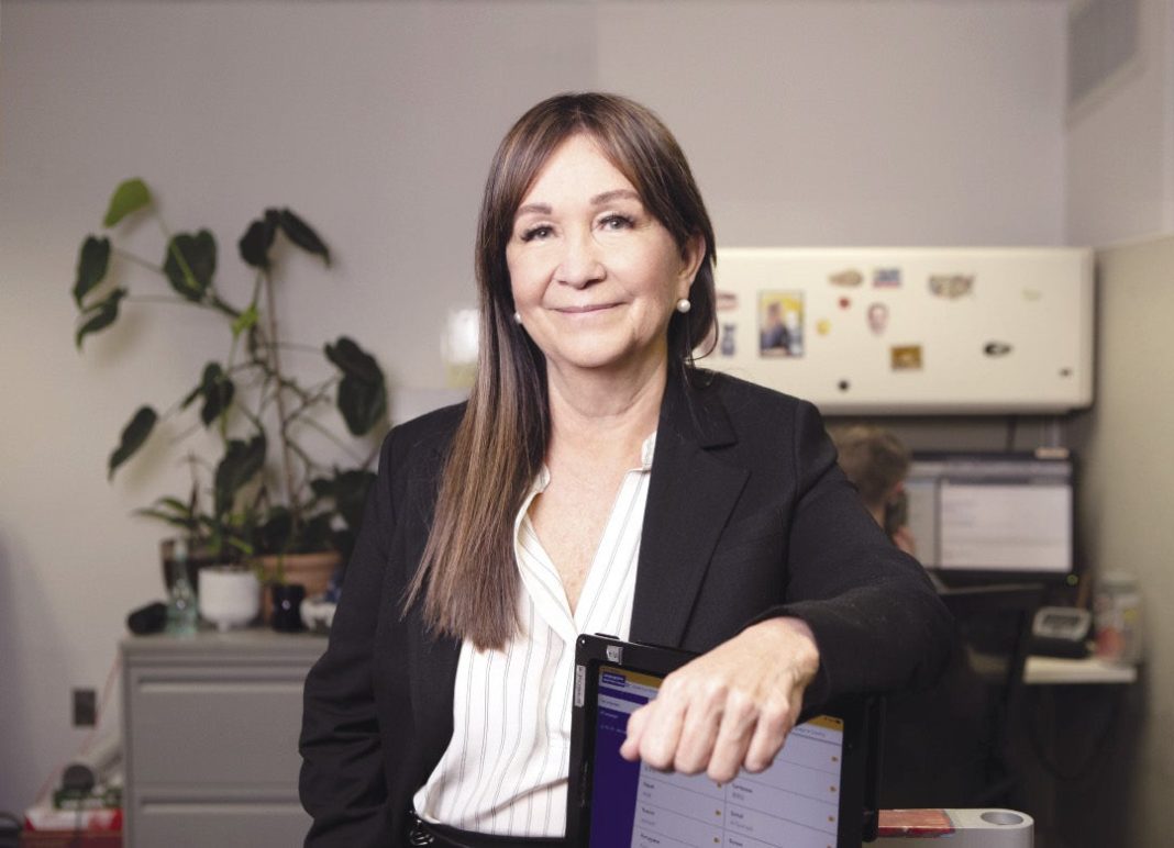 A woman in a black blazer and white shirt stands behind a computer screen.