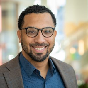 A man with dark hair, a beard, and black glasses wears a grey striped suit with a blue dotted button down.
