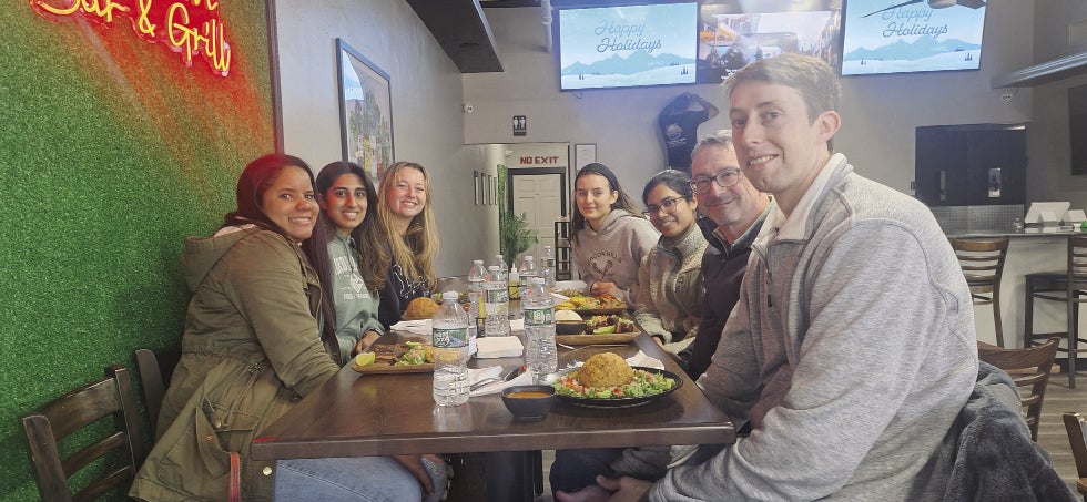 A group of seven people sit around a rectangle high top chair in a restaurant with plates of food in front of them.