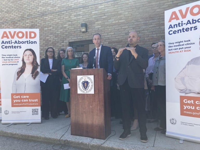 A man stands behind a wooden podium on a concrete sidewalk with 9 others around him in between signs reading 