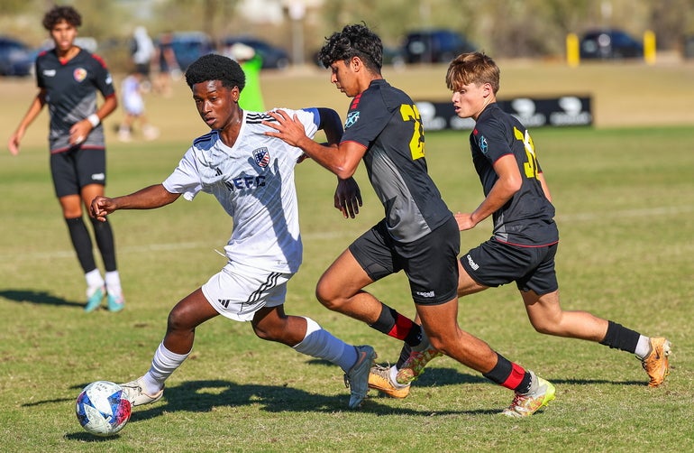 A soccer player in a white uniform dribbles a ball at his feet while two defenders in black uniforms chase him