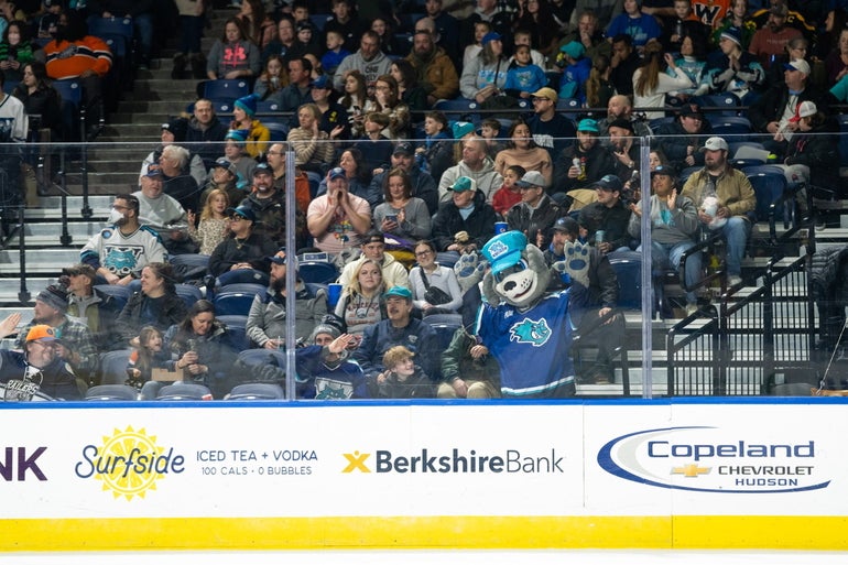 A dog mascot pushes itself up against the glass of a hockey rink, with a crowd behind it