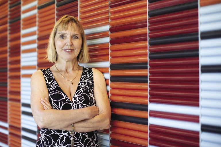 A woman in a black floral dress leans against a multi-colored wall made of rounded wood panels