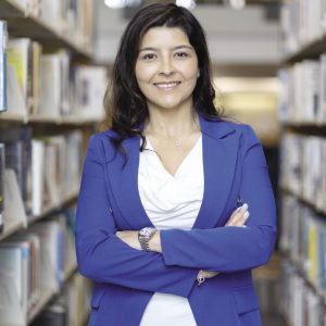 A woman with dark brown hair wears a blue suit jacket and white blouse with folded arms and a silver watch in a library aisle.