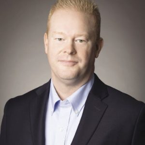 A man with short blonde hair wears a black suit jacket and light blue button up shirt.