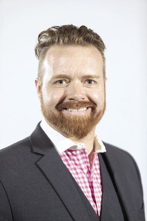 A man with red hair a red beard wears a dark grey suit jacket and white and pink checkered button down.