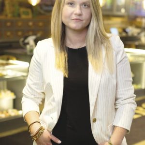 A woman with blond hair wearing a a black shirt and white jacket stands in front of a sign, which says "Caroline's Cannabis".