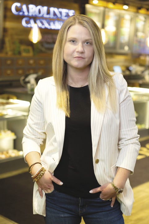 A woman with blond hair wearing a a black shirt and white jacket stands in front of a sign, which says 