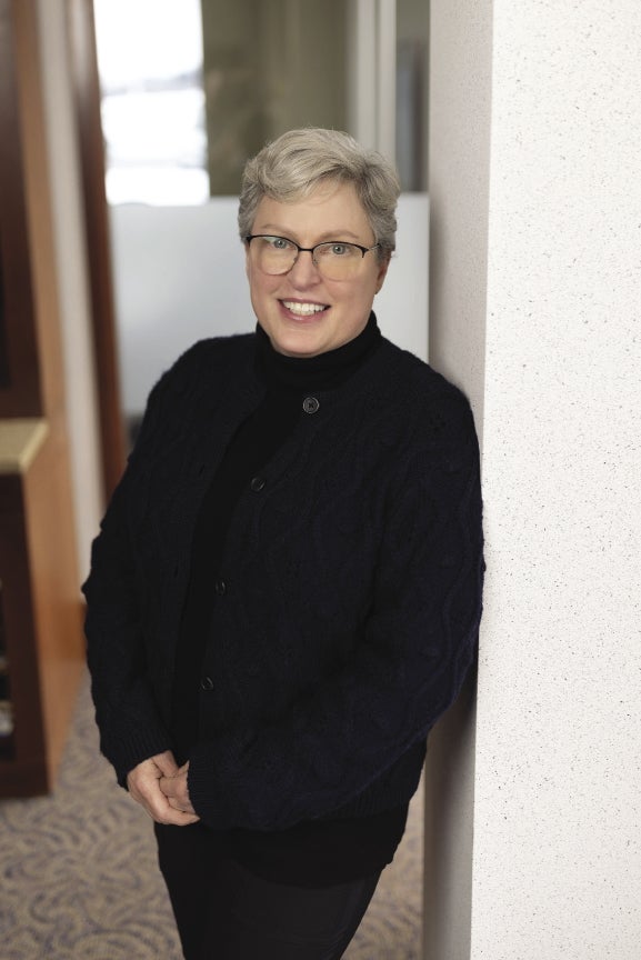 A woman with short white hair wears a black turtleneck and black pants.