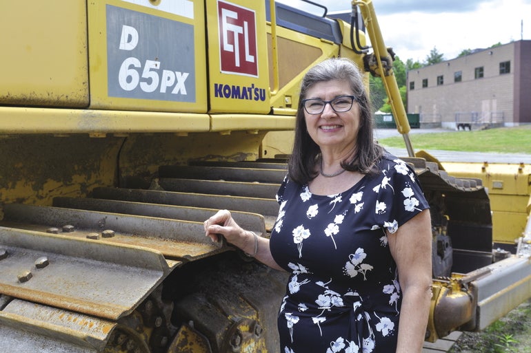 A woman in a black dress with a white, floral pattern stands with her hand on a large yellow front loader.