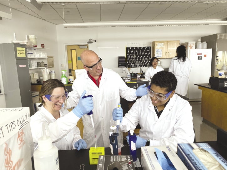 Two MassBioEd aprentices receive instruction from a lab teacher. They are in a classroom setting using pipettes.