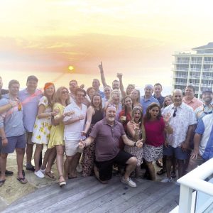 A large group of people stand in front of a setting sun at a resort.