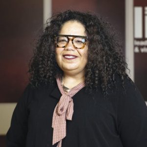 A woman with dark curly hair wears a black top and black and tortoise glasses.