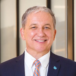 Edward Manzi, wearing a blue and orange rep tie with a suit coat and white shirt, smiles at the camera.