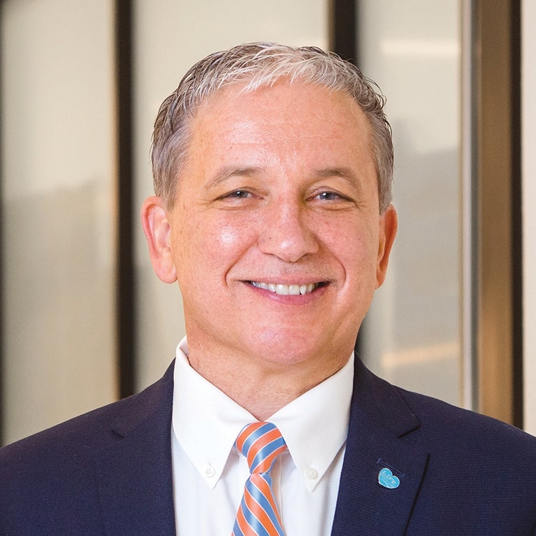 Edward Manzi, wearing a blue and orange rep tie with a suit coat and white shirt, smiles at the camera.