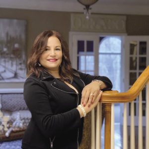 A women with brown hair wears a black jacket and skirt, leaning against a stair railing