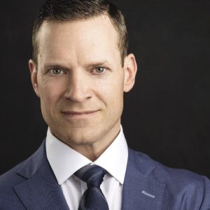 A man in a blue suit, white shirt, and blue tie stares intently at the camera.