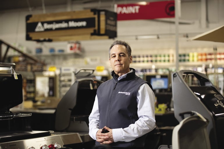 A man in a light blue shirt and grey vest stands with hands together looking at the camera in a hardware store with grills around him.