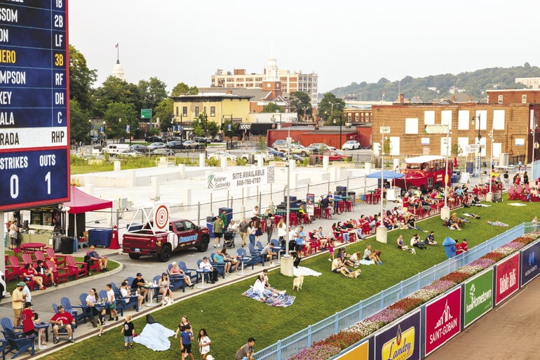 People sit on a grass bern in the left field area of a minor league ballpark with an undeveloped commerical site fenced off behind them.