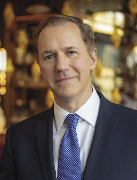 A man with light brown hair wears a dark blue suit jacket, white button down, and blue patterned tie.