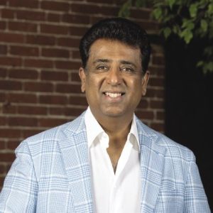 A man with short dark hair wears a light blue and white plaid suit jacket and a white button down.