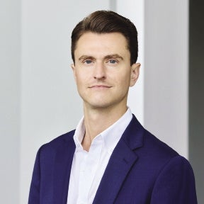 A headshot of a man with brown hair wearing a dark blue suit jacket and white button down