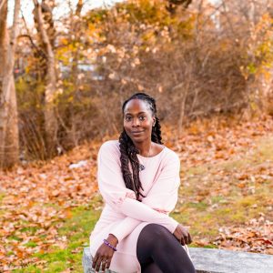 A woman with long dark brown hair wears a light pink dress and dark tights sitting on a grey stone bench in front of fall foliage.