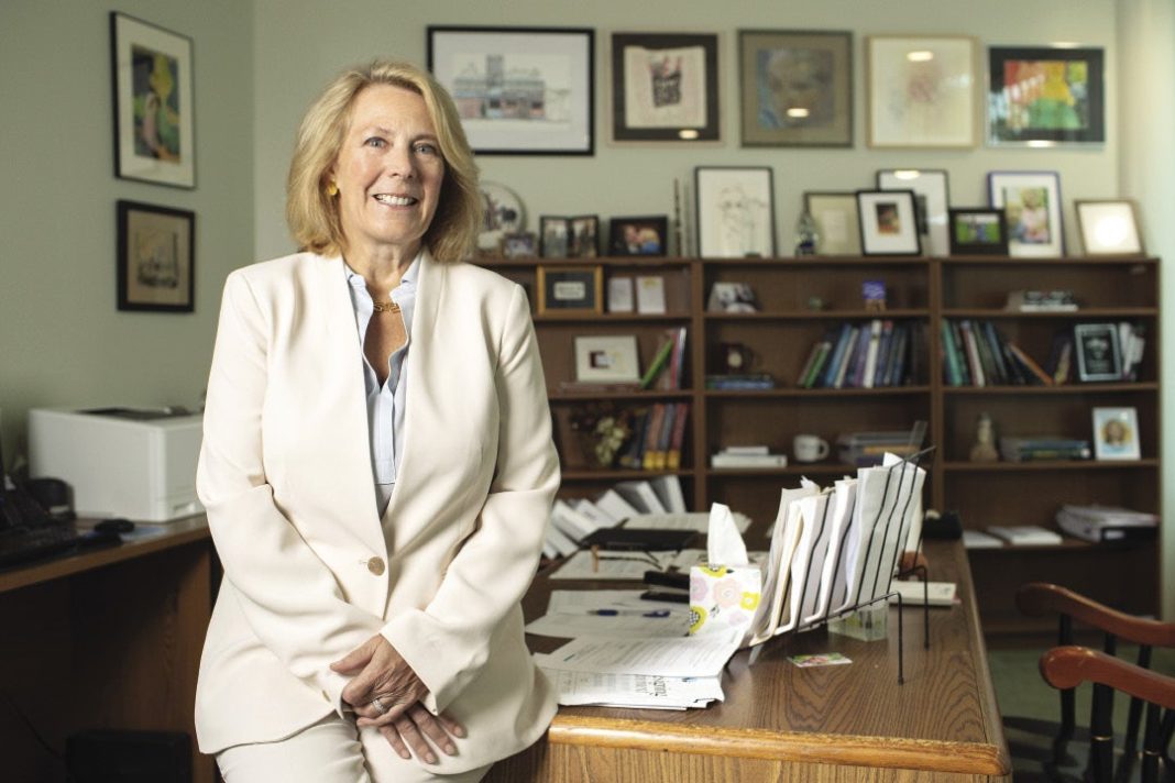 A woman in a white suit leans on her desk in her office.