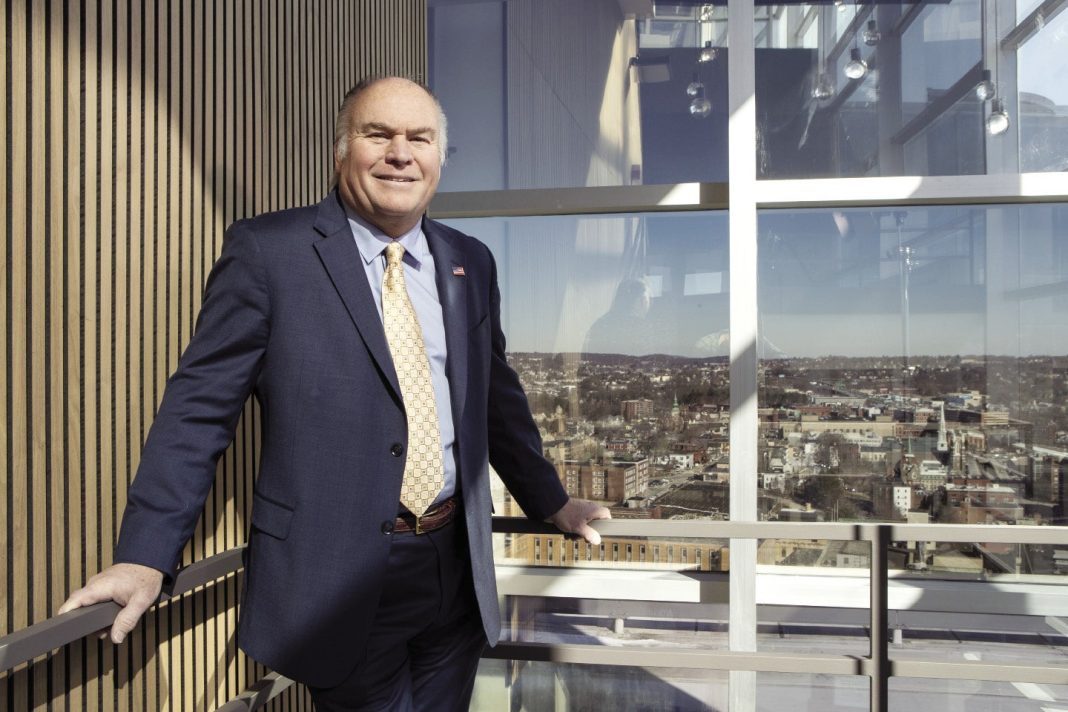 A man in a suit stands in front of a window overlooking the Worcester skyline