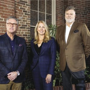Three people in business attire stand in front of a brick background