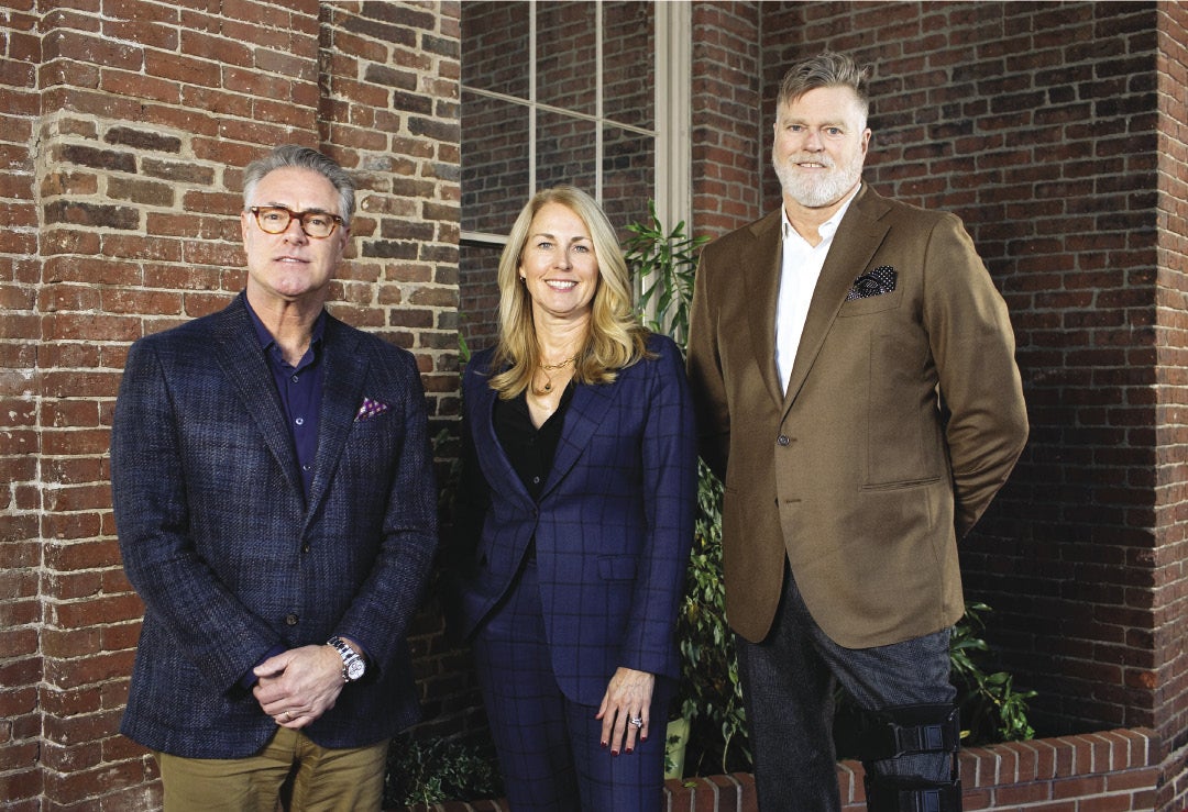 Three people in business attire stand in front of a brick background