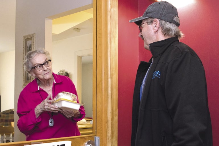 A woman with grey hair and glasses wearing a pink button down holds a couple cartons of food as she looks through a door window at a man with grey hair wearing a grey baseball cap and a black zip up jacket.