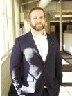 A man with a beard wears a dark blue suit jacket and white button down with his hands in his pockets.