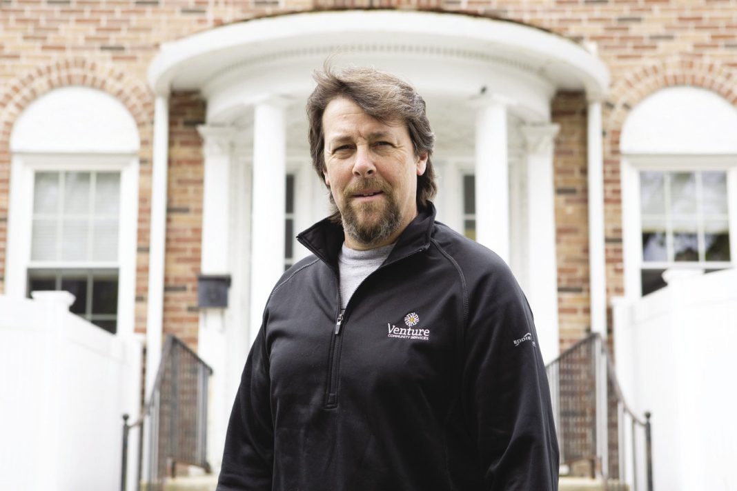 A man with brown hair and a goatee wears a black zip-up jacket in front of a brick building.