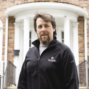 A man with brown hair and a goatee wears a black zip-up jacket in front of a brick building.