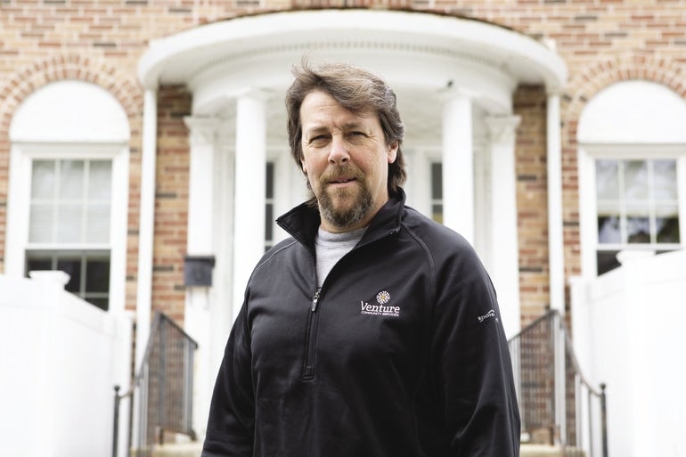 A man with brown hair and a goatee wears a black zip-up jacket in front of a brick building.