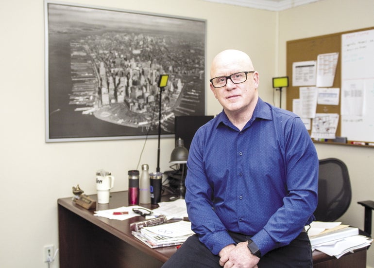 A man wearing a blue shirt and glasses sits against his desk.