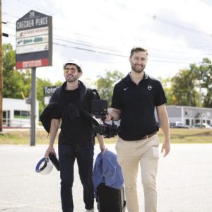 two men carrying equipment walk toward the camera in a parking lot