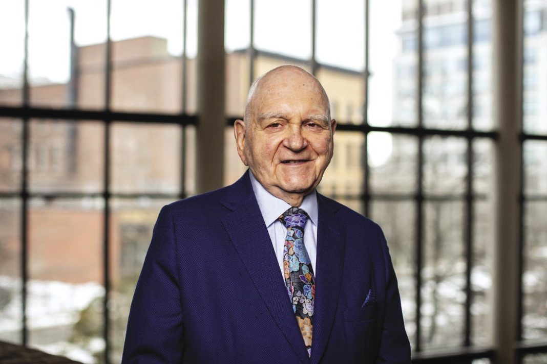A man in a suit in front of large windows with cityscape behind