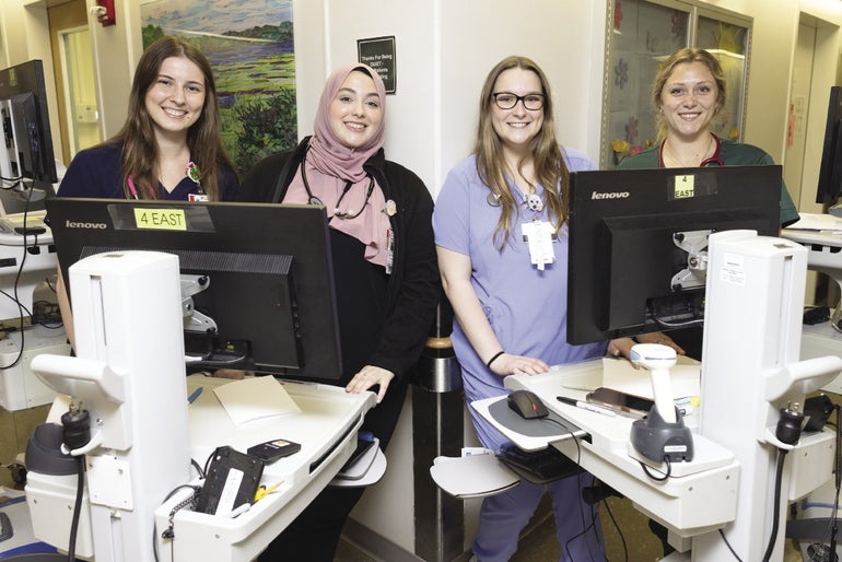 Four woman nurses stand behind rolling carts with computer screens against a cornered wall in a hospital.
