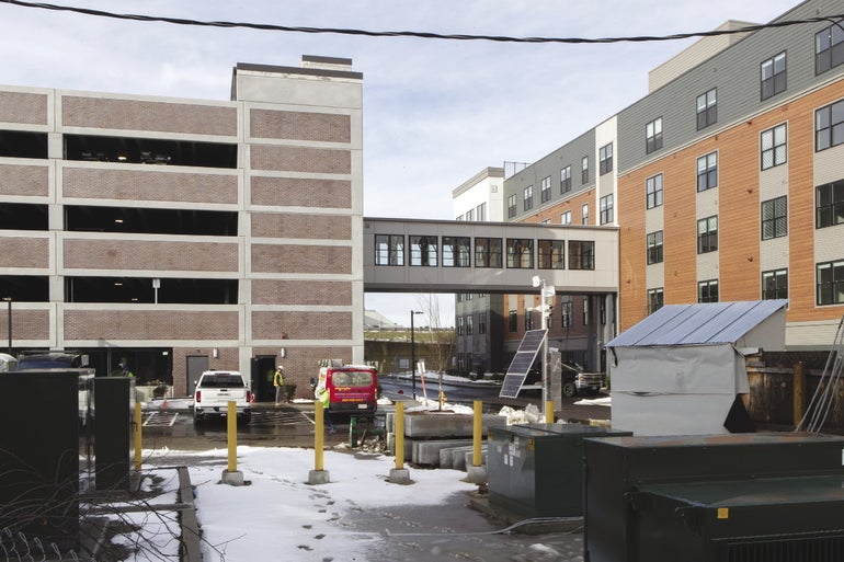 A five story parking garage sits across a small street from a large apartment building, with a footbridge connecting the two structures.