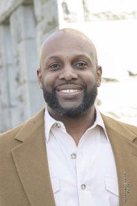 A man with a black beard wears a tan suit jacket and white button down