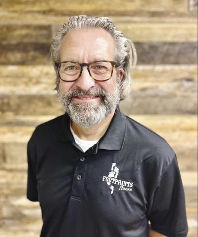 Sean Donohoe stands with his hands behind his back, looking at the camera with a smile peaking out through his gray beard and his blue eyes glistening behind his glasses.