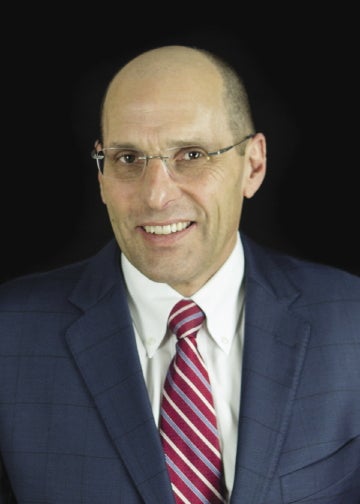 A headshot of a man wearing rimless glasses, a dark blue suit jacket, white button down, and a red and white striped tie.