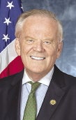 A man with white hair wears a black suit jacket, a blue and white button down, and a green tie.